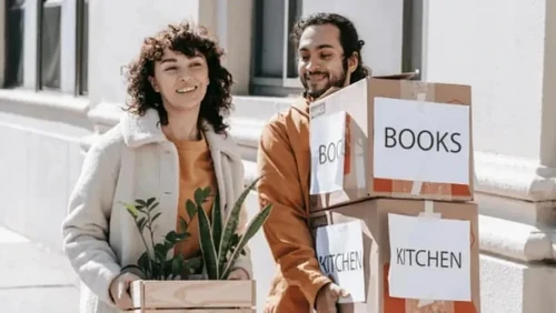 Young couple walking with boxes of books, kitchen supplies, and plants